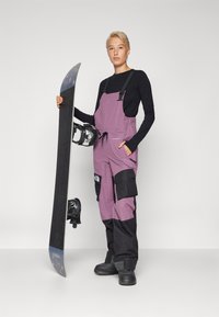 Lumelauguja, kes kannab musta pika varrukaga paela ja lillaka-musta kombinesooni, hoiab lumelauda koos kinnitustega, demonstreerides taskuid ja reguleeritavaid rihmasid.