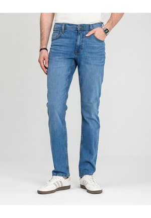 Mężczyzna w jasnoniebieskich, dopasowanych dżinsach, białych sneakersach z szarymi paskami, zegarkiem na lewej ręce oraz bransoletką na prawej, stojący na tle szarego tła.