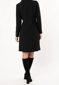 Donna che indossa un cappotto nero al ginocchio e stivali neri con tacco al ginocchio, in piedi e di spalle su uno sfondo bianco.