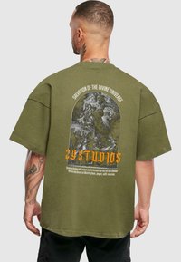 Olivengrøn oversized T-shirt med et grafisk print på ryggen, der indeholder en illustration og tekst i orange og hvid. Korte ærmer.