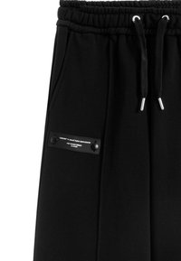 Pantalones deportivos negros con cintura elástica, cordones, bolsillos laterales y un parche de marca con botones a presión en la pierna izquierda.