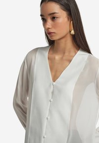 Blusa bianca semi-trasparente con scollatura a V, dotata di chiusura frontale con bottoni e sottili cuciture verticali. Le maniche lunghe e fluide aggiungono eleganza.