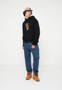 Jovem vestido com um boné bege, uma sweatshirt preta com um gráfico de cavalo, jeans azuis e botas de trabalho castanhas, posicionado contra um fundo branco.