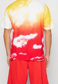 Koszulka z gradientem od żółtego do czerwonego z wzorami chmur. Krótkie rękawy, wykonana z miękkiego materiału. Zestawiona z czerwonymi szortami.