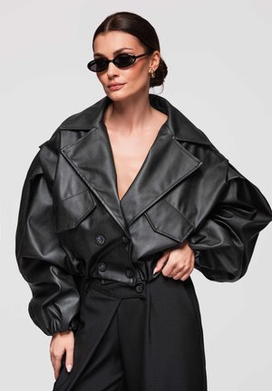 Frau trägt übergroße schwarze Lederjacke und schmale schwarze Sonnenbrille, posiert mit Hand in der Hüfte vor einfarbigem Hintergrund.