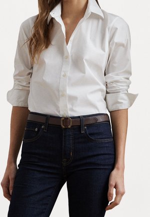 Frau trägt ein weißes, geknöpftes Hemd mit hochgekrempelten Ärmeln, das in dunkelblaue Jeans gesteckt ist, und einen braunen Gürtel mit goldener Schnalle.