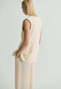 Top sin mangas de color beige con un corte holgado, que presenta un cuello alto y aberturas laterales, combinado con pantalones anchos a juego. Textura suave.