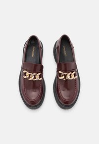 Pár vínových lakovaných loafers s robustními černými podrážkami a výrazným zlatým řetízkem přes přední část.