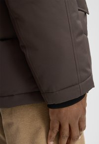 La manica della giacca a vento marrone presenta righe orizzontali, cuciture a sbalzo e un polsino a maglia nera. La mano è appoggiata su un tessuto testurizzato color beige chiaro.