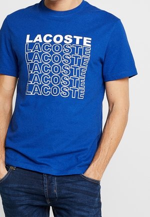 Homme portant un t-shirt bleu vif à manches courtes avec le mot "LACOSTE" répété six fois en texte blanc sur le devant, associé à un jean bleu foncé.