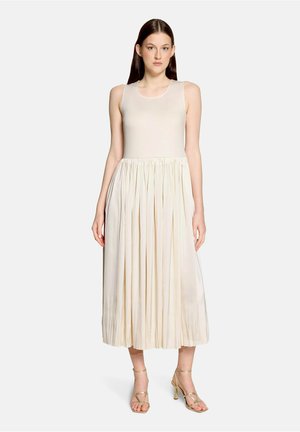 Kobieta z prostymi ciemnymi włosami, ubrana w kremowy top bez rękawów oraz sukienkę z plisowaną midi spódnicą i złote sandały na obcasie z paskami.