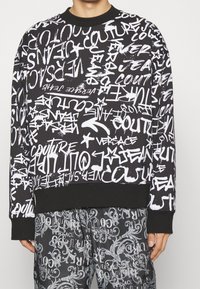 Černá mikina s bílým graffiti-style textem a vzory. Obsahuje vroubkovaný kulatý výstřih a manžety. V kombinaci s kalhotami se shodným vzorem.