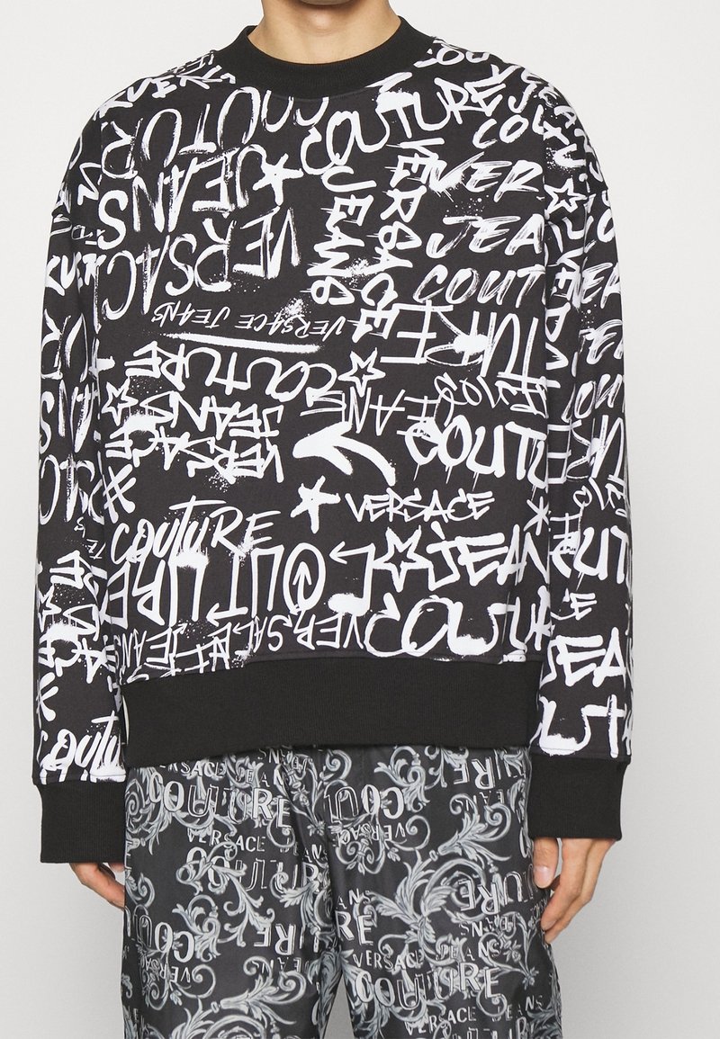 Černá mikina s bílým graffiti-style textem a vzory. Obsahuje vroubkovaný kulatý výstřih a manžety. V kombinaci s kalhotami se shodným vzorem.