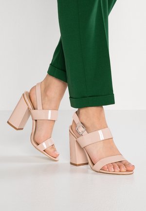 Feet wearing nude patent leather block heel sandals with ankle straps, paired with rolled-up green pants on a white floor.