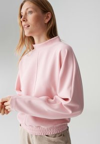 Roze trui met een hoge kraag, wijde mouwen en een geplooide zoom. Gemaakt van zachte stof met een gladde textuur en subtiele details.