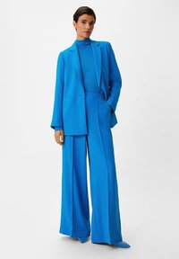 Blauw op maat gemaakte pak met wijde broekspijpen en een nauwsluitende coltrui. Bevat een blazer met ontsloten revers en een gladde stoftextuur.