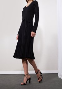 Robe noire à manches longues avec un corsage ajusté, une jupe plissée et une ceinture nouée à la taille ; portée avec des sandales à talons hauts noires à lanières.