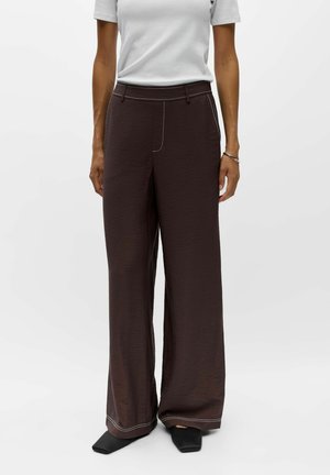 Personne portant un pantalon large marron foncé avec des coutures blanches, des chaussures noires à bout carré et un haut gris clair à manches courtes, debout sur un fond blanc.