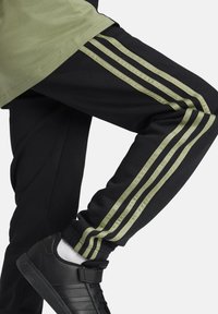 Pantalones de chándal negros con tres franjas verde oliva a lo largo de los costados, usados con unas zapatillas negras y calcetines blancos. Material de mezcla de algodón.