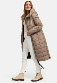 Lange gewatteerde jas in beige, met een capuchon, ritssluiting en een gestikt patroon. Gecombineerd met witte broeken en beige veterschoenen.
