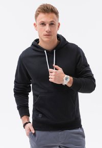 Felpa nera con cappuccio e lacci, tasca frontale e polsini a coste. Indossata con pantaloni grigi; presenta un orologio bianco al polso.