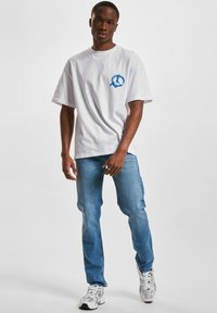 Hvit bomulls t-skjorte med blå fredstegn grafikk på venstre bryst, kombinert med lyseblå jeans og hvite joggesko.