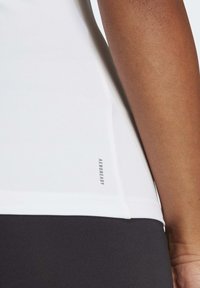 Top atletico bianco con una superficie liscia, caratterizzato da un sottile etichetta AEROREADY sul lato. Combinato con un tessuto nero visibile sotto.