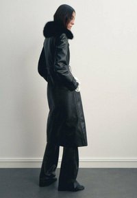 Cappotto in pelle nera con colletto in pelliccia, maniche lunghe e tasche laterali. Indossato sopra pantaloni neri larghi su una figura contro uno sfondo neutro.