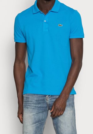 Homme portant un polo à manches courtes bleu vif avec un petit logo crocodile sur la poitrine, associé à un jean bleu clair délavé.