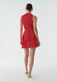 Punainen hihaton mekko, jossa kerroksellinen hame ja teksturoitu kangas. Liehuva helma ja istuva vyötärö, täydentyy yksinkertaisilla kengillä.