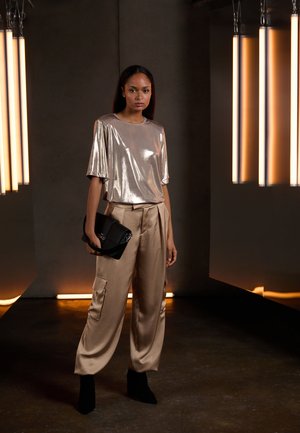 Metallic sølv kortærmet top med løs pasform, parret med beige satin cargo bukser. Sort håndtaske og sorte ankelstøvler. Varm omgivende belysning.