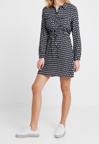 Robe noire à pois blancs, taille cintrée, manches longues, fermeture à boutons à l'avant, deux poches sur la poitrine, associée à des baskets blanches.