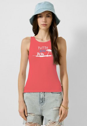 Femme portant un débardeur corail avec un motif de chaise de plage et parasol, un short en jean clair déchiré et un chapeau bob bleu clair, debout devant un fond uni.