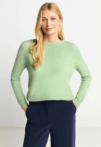 Hellgrüner Wollpullover mit Rundhalsausschnitt und langen Ärmeln, der eine weiche Textur und gerippte Manschetten aufweist, kombiniert mit navyfarbenen Hosen.