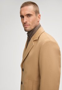 Cappotto in misto lana beige con design doppiopetto, rever a scalino, bottoni scuri e vestibilità sartoriale sopra una giacca marrone a colletto alto.
