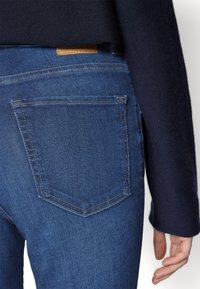 Jeans in denim blu con un design aderente, caratterizzati da una tasca posteriore pulita, una toppa in pelle color tannico e dettagli di cucitura sottili.