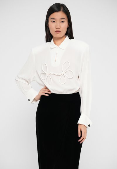 White long-sleeve shirt with a collar, featuring a textured decorative detail resembling loops at the chest. Paired with a black skirt.