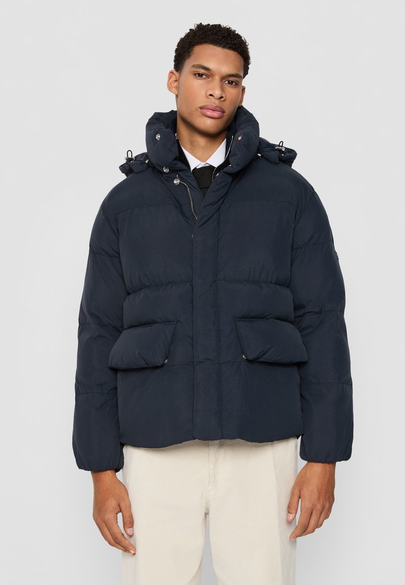 Doudoune marine avec un col haut, fermeture éclair à l'avant et deux poches latérales. Présente une texture matelassée et des poignets élastiqués pour plus de chaleur.