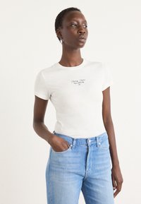 T-shirt bianca a maniche corte con la scritta "Tommy Jeans New York City" stampata in nero, abbinata a jeans di denim blu chiaro. Il modello ha una postura rilassata.