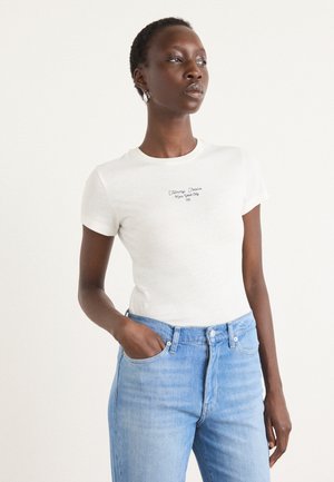 Īsa piedurkņu balta T-kreklu ar melnu uzrakstu "Tommy Jeans New York City", apvienots ar gaiši ziliem džinsiem. Modelis atrodas relaksētā pozā.
