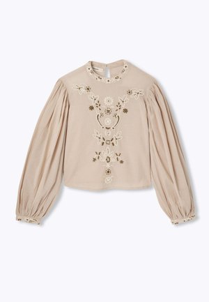 Blouse beige avec des motifs floraux brodés en crème et vert. Dotée de manches longues bouffantes et d'une ouverture en forme de clé au dos.