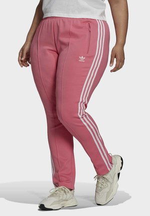 Osoba w różowych spodniach dresowych Adidas z białymi paskami i kieszeniami na zamek, w połączeniu z białymi trampkami na jednolitym tle.