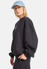 Felpa nera oversized con polsini a costine, con un piccolo logo sul petto. Abbinata a un cappellino in denim blu chiaro.