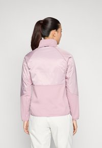 Giacca sportiva di un delicato rosa chiaro con colletto alto, texture morbida, maniche raglan e design aderente, adatta per l'abbigliamento attivo.