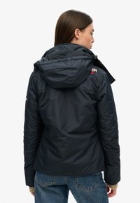 Veste isolante bleu marine avec une capuche, présentant une finition texturée et un accent logo près du col. La vue de dos montre un design ajusté.