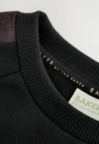 Melnā džemperis ar ribotu apkakli, ar teksturētu pārklājumu un zelta etiķeti ar uzrakstu "BAKER by Ted Baker." Iekšpuse ir mīksta.