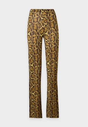 Kvinde med langt sort hår iført en gul og brun slangeprint off-shoulder tætsiddende jumpsuit og sorte sko, stående på en hvid baggrund.