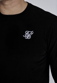 Melnas garām piedurknēm T-krekls ar mīkstu tekstūru un apaļu kakla izgriezumu, ar baltu izšūtu logotipu kreisajā krūtīs.