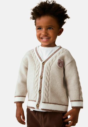 Lächelndes Kind, das eine cremefarbene Strickjacke mit Zopfmuster, braunem Besatz und einem Teddybär-Patch auf der Brust trägt, steht vor einem schlichten Hintergrund.
