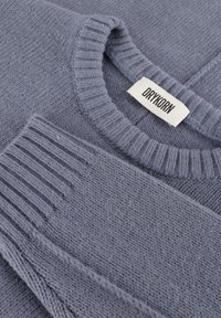 Grauer Strickpullover mit einem gerippten Rundhalsausschnitt und Ärmelbündchen, ausgestattet mit einem strukturierten Muster und einem etikettierten Tag. Weiches, wool-ähnliches Material.